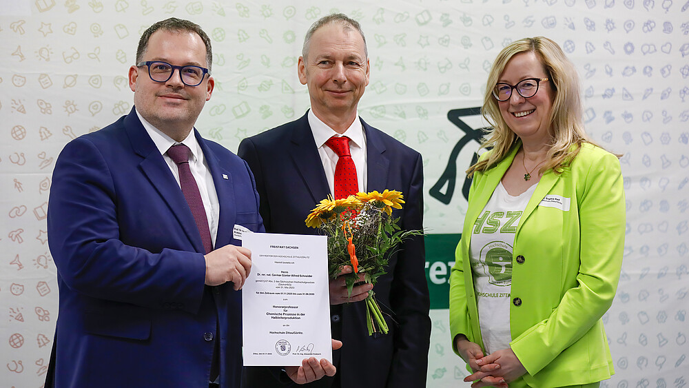 Rektor, Herr Schneider und die Prorektorin stehen vor einer Pressewand und lächeln in die Kamera.