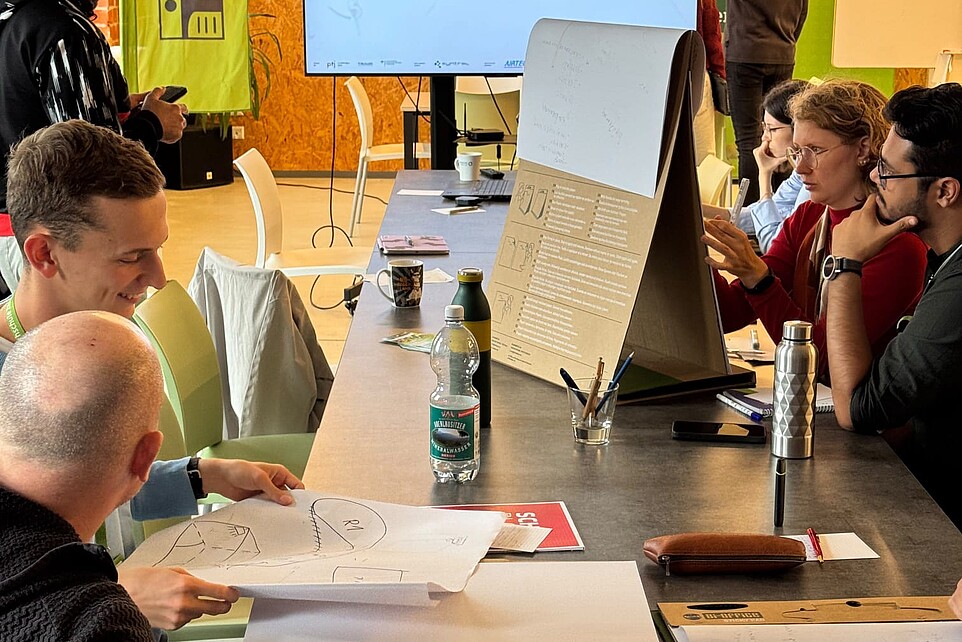 In einem Seminarraum sitzen Schüler an einem Tisch und schauen auf einem Bildschirm.