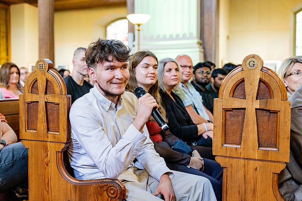 Ein Erstsemester sitzt in einer Kirchenbank der Johanniskirche und spricht mit einem Handmikrofon.