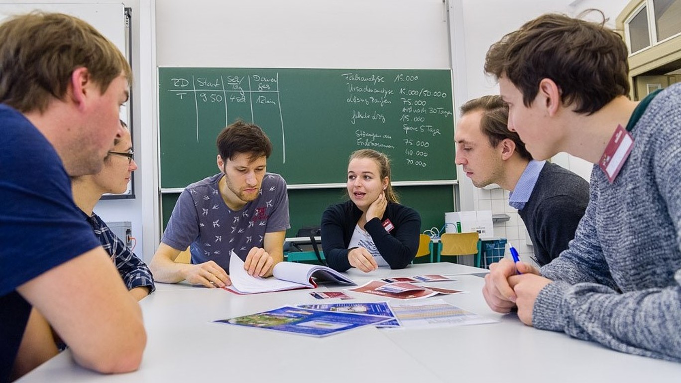 Am Planspiel Teilnehmende sitzen am Tisch, blättern in Unterlagen und unterhalten sich.