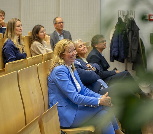 Die Sitzreihen sind mit Publikum besetzt, dass den Blick nach vorn richtet.