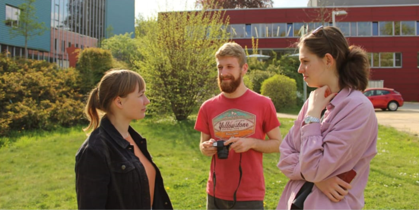drei Studierende besprechen sich, im Hintergrund die Mensa