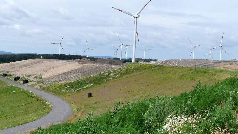 Abfallwirtschaft praktisch. Außenansicht auf die Deponie Wetro. Begrünte Hügel und abgetragene Flächen im Vordergrund, Windräder im Hintergrund.