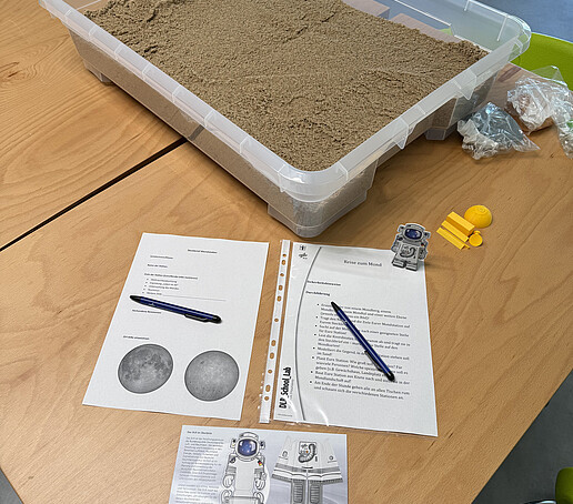 Eine Sandbox und Ausdrucke zum Mond liegen auf einer Holztischplatte.