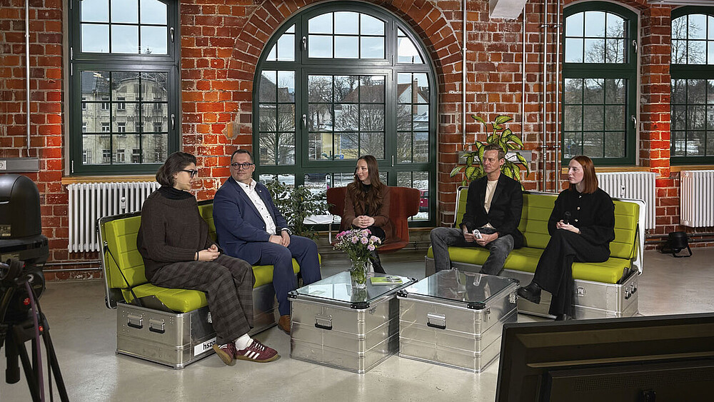 Morning Show-Teilnehmende sitzen auf grünen Sofas im TV-Studio.