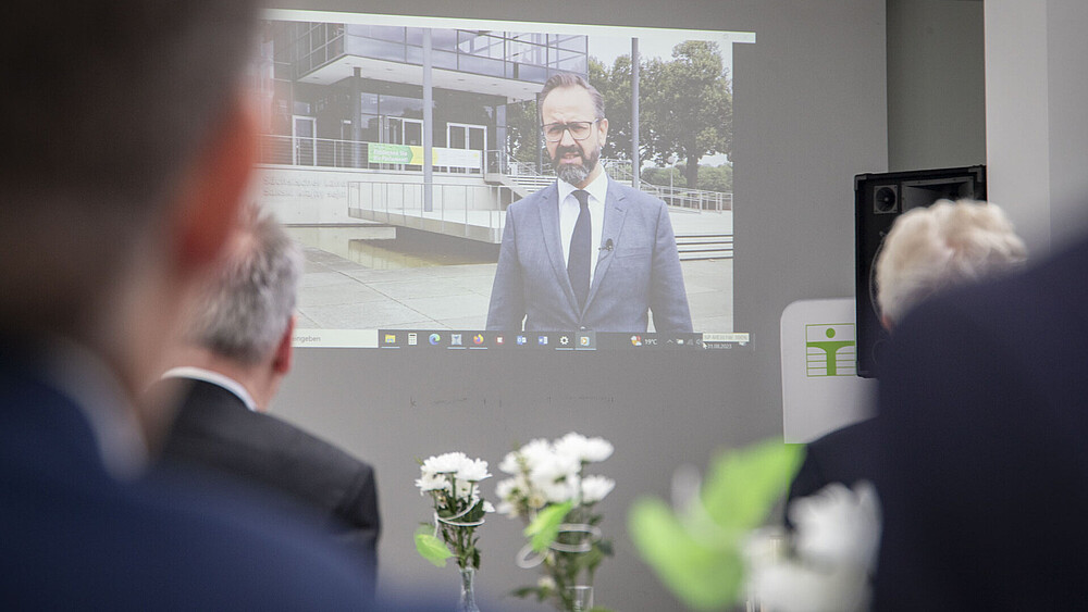Videoübertragung über Beamer an einer Wand mit Staatsminister Gemkow. Gäste schauen sich seine Rede an.