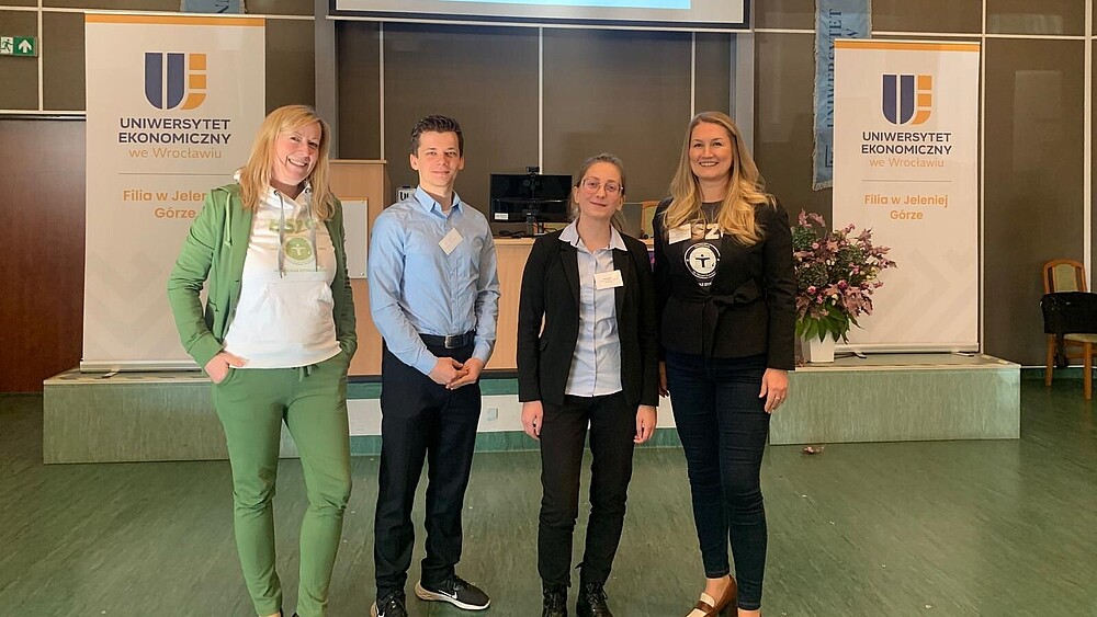 Vier Vertretende der HSZG stehen in einer mit Roll-ups geschmückten Halle nebeneinander und lächeln in die Kamera.