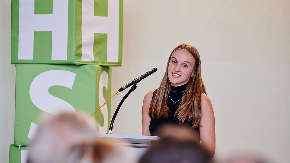 Carla Wagner steht auf der Bühne am Rednerpult und spricht ins Mikrofon.