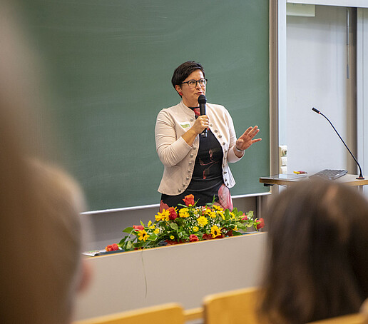 Eine Frau spricht in ein Mikrofon vor einer grünen Tafel im Hörsaal.