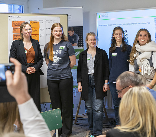 Eine Gruppe aus vier Frauen stehen vor vollbesetzten Stuhlreihen. Im Hintergrund steht eine Pinnwand und an der Wand hängt ein großer Bildschirm mit einer Präsentation.