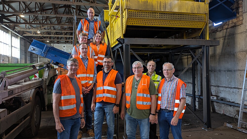Eine Gruppe von neun Personen steht in einer Industriehalle vor einer großen gelben Sortier- oder Recyclinganlage. Alle tragen orange Sicherheitswesten und blicken in die Kamera.