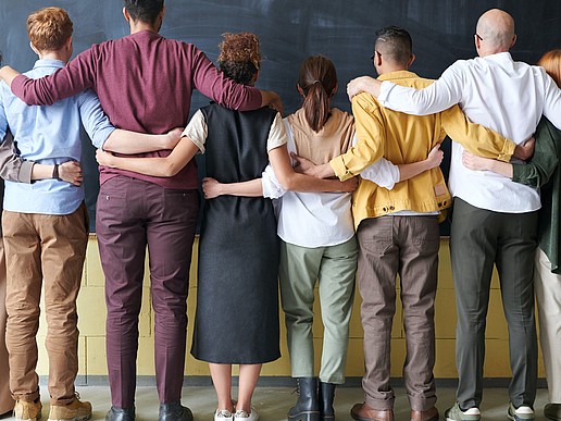 Eine Reihe von junger Menschen unterschiedlicher ethnischer Herkunft stehen in einer Reihe, den Rücken zur Kamera gedreht, mit Blick auf eine Tafel. Sie legen sich gegenseitig die Hände und Arme auf den jeweiligen Danebenstehenden.