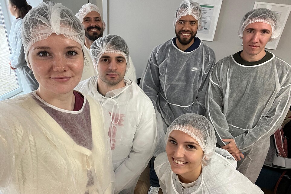 Eine Gruppe Studierender in Schutzanzügen und Kopfbedeckungen lächeln in die Kamera.