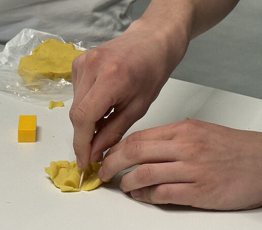 Zwei Hände bearbeiten ein Stück gelbe Knete mit einem Schneidewerkzeug.